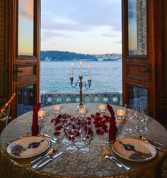 a table set for two with candles and flowers in front of an open window overlooking the water