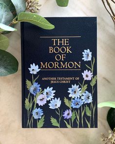 the book of mormon with blue flowers and green leaves on a marble table next to it