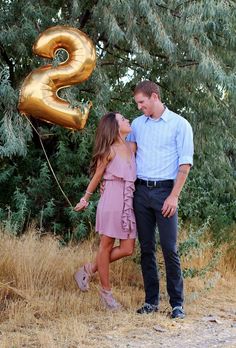 a man and woman standing next to each other in front of trees with gold balloons