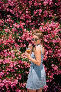 a woman standing in front of pink flowers drinking from a wine glass and looking at her cell phone