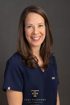 a woman wearing a blue scrub top smiles at the camera while standing in front of a gray background