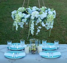 a table set with blue and white plates, napkins, candles and flowers hanging from the ceiling