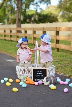 two small children sitting in a basket on the ground with easter eggs and carrots