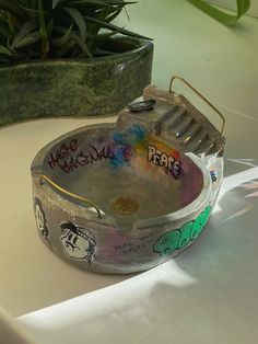 a metal bowl with graffiti on it sitting on top of a table next to a potted plant