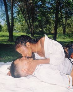 a man and woman laying on top of a blanket in the grass next to trees