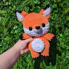 a hand holding an orange and black knitted toy in front of some green bushes