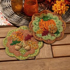 two decorative fall coasters sitting on top of a wooden table next to a basket filled with flowers