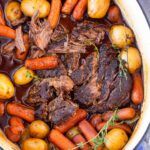 pot roast with carrots, potatoes and other vegetables in a white bowl on top of a blue table cloth