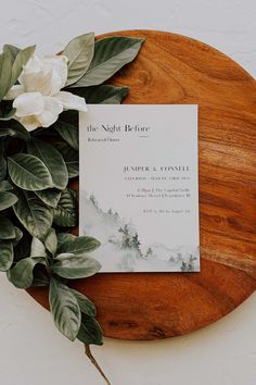 the wedding stationery is laid out on a wooden platter with flowers and greenery