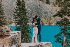 a man and woman standing next to each other on top of a cliff near water