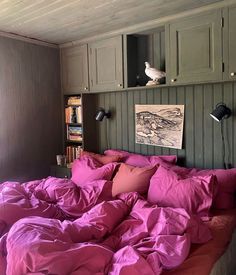 an unmade bed with pink sheets and pillows in front of a book shelf filled with books