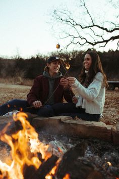 a man and woman sitting next to each other in front of a campfire holding hands