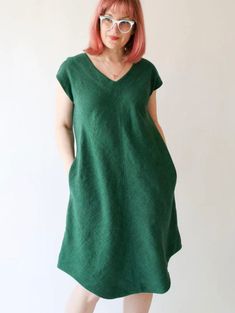 a woman with pink hair and glasses standing in front of a white wall wearing a green dress