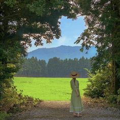 a woman in a dress and hat standing on a dirt road with trees around her