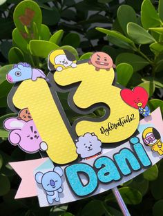 a birthday cake topper with the number 13 and cartoon characters on it, sitting in front of some green leaves