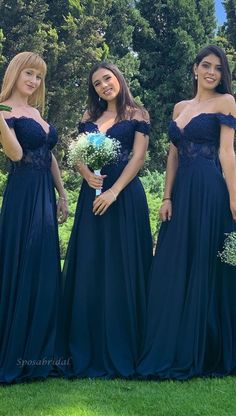 three women in long dresses standing on grass with trees in the background and one woman holding a bouquet