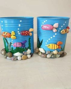 two blue tin cans with fish painted on them sitting next to rocks and pebbles in the water