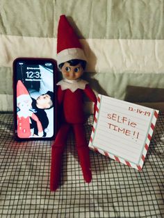 an elf is sitting next to a cell phone and sign that says selfie time