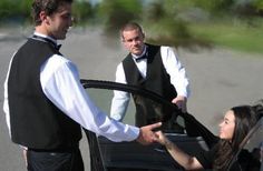 two men and a woman standing next to a car with their hands on the steering wheel