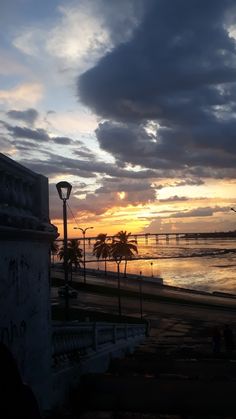 the sun is setting over the ocean and people are walking on the sidewalk near the beach