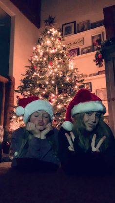 two women wearing santa hats sitting in front of a christmas tree with lights on it