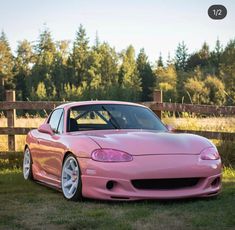 a pink sports car parked on the grass in front of a wooden fence and trees