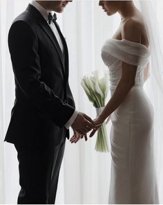 a bride and groom standing next to each other in front of a window holding hands