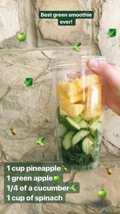 a hand holding a cup filled with cucumber and pineapple slices in front of a stone wall