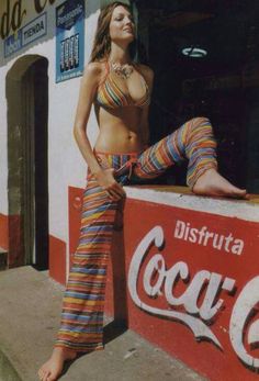 a woman leaning on a coca - cola sign in front of a building with her legs crossed