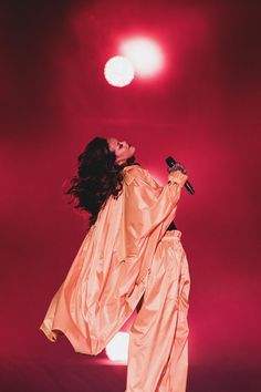 a woman in an orange outfit holding a microphone and singing into the air with her arms outstretched