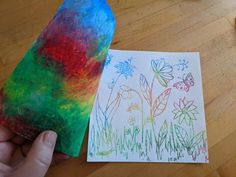 a child's hand holding a piece of paper next to an art project with flowers and butterflies on it