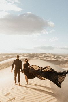 a man and woman walking in the sand with a long black dress on their back