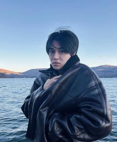 a young man standing on top of a boat near the ocean wearing a black leather jacket