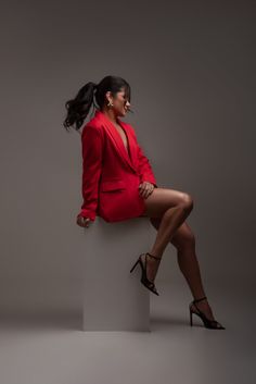 a woman in a red suit sitting on top of a white block with her legs crossed