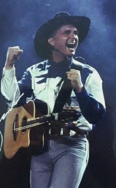 a man holding a guitar and singing into a microphone with his right hand up in the air