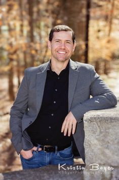 a smiling man leaning against a stone wall in the woods with his hands on his hips