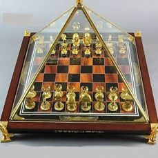 a glass and wood chess board with golden figures on it's sides, in front of a gray background