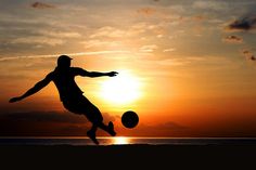 a man kicking a soccer ball on top of a sandy beach at sun set in the background