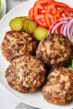 some meat patties are on a white plate with tomatoes, cucumbers and pickles