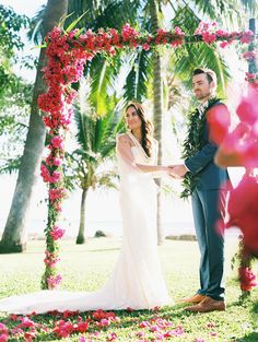 a bride and groom holding hands under an archway with pink flowers on the grass in front of palm trees