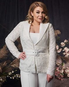 a woman standing in front of a floral arrangement wearing a white jacket and pants with lace detailing