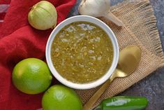 a white bowl filled with green liquid next to two limes and an onion on a red towel