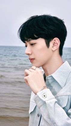 a young man standing in front of the ocean with his hand on his chin and looking off into the distance