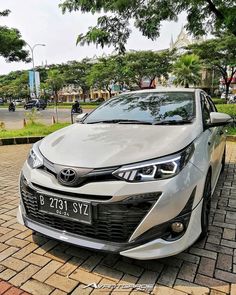 the front end of a silver car parked on a brick road near trees and bushes