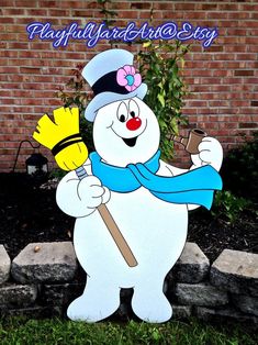 a snowman holding a broom standing in front of a brick wall and grass area