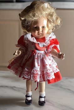 This 14-inch Shirley Temple doll is a delightful representation of the iconic child star, dressed in a charming red dress with a white apron adorned with red hearts. The apron features red bows, and there is a red bow at her belly, adding to the sweetness of her outfit. Her dress is trimmed with lace around the collar and the short puffy sleeves, enhancing her classic, adorable look. She proudly wears a pin that says "My Friend Shirley Temple" and has Shirley Temple's signature curls, crowned with a big white bow in her hair. Her expressive green sleepy eyes are made of glass and can open and close, capturing the lively spirit of Shirley Temple. She has a radiant smile with red lips, showing her teeth and dimples. While this Shirley Temple doll is a cherished collectible, please note that Child Star, White Apron, Sleepy Eyes, Shirley Temple, Red Hearts, White Bow, Puffy Sleeves, Collector Dolls, Red Bow