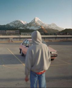 a person in a hoodie walking towards a car parked in a parking lot with mountains in the background