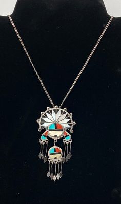 Beautiful Zuni handmade pendant necklace is made of sterling silver, and has multi-stone inlay with sterling silver "raindrops" hanging down. The pendant has 2 Zuni Sun Face Kachinas; the one on the top has a headdress and the bottom one is hanging down vertically from the top one. Each of the Sun Face Kachinas have sterling silver "raindrops" hanging down from them. There are 5 hanging down from the bottom face and 6 hanging down from the top one (3 on each side). The pendant has multi-stone inlay of turquoise, mother of pearl, jet, and spiny oyster. The chain that the pendant is currently on is stamped 835 which is 83.5% pure silver, but it can be replaced and used with just about any thin chain if desired.  Condition: Excellent! Minor wear consistent with age and use  Artist: Unknown/Un Handmade Pendant Necklace, Sun Face, Stone Inlay, Spiny Oyster, Sterling Silver Necklace Pendants, Long Pendant, Handmade Pendant, Multi Stone, Silver Pendant Necklace