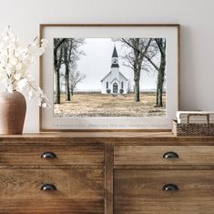 a framed photograph of a white church in the middle of a field next to a dresser