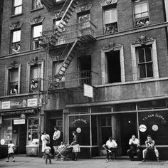 an old black and white photo of people sitting on the sidewalk in front of a building